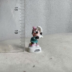 May include: A small Minnie Mouse figurine, featuring a pink bow, black ears, and a teal dress. The figurine is approximately 1 inch tall and stands on a white base with the text "Minnie Disney".