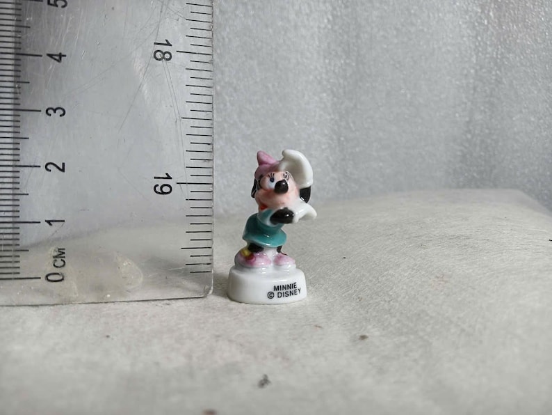 May include: A small Minnie Mouse figurine, featuring a pink bow, black ears, and a teal dress. The figurine is approximately 1 inch tall and stands on a white base with the text "Minnie Disney".