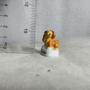 May include: A small, ceramic dog figurine in shades of orange and brown, standing on a white base. The figurine is approximately 1 inch tall. The base has the word "DISNEY" printed on it. The dog has a happy expression.