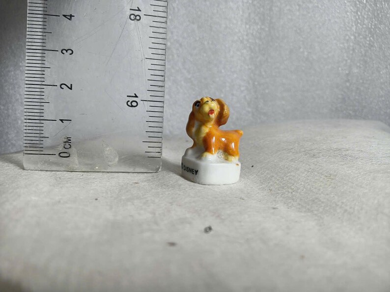 May include: A small, ceramic dog figurine in shades of orange and brown, standing on a white base. The figurine is approximately 1 inch tall. The base has the word "DISNEY" printed on it. The dog has a happy expression.