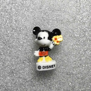 Op de afbeelding: Een klein keramieken Mickey Mouse beeldje. Mickey draagt een zwart jasje, een rode korte broek en gele schoenen. Hij houdt een geel bloemenboeket vast. De basis van het beeldje is wit met het woord "DISNEY" erop.