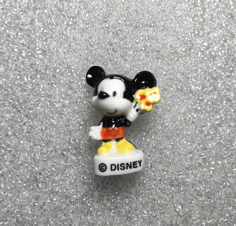 Op de afbeelding: Een klein keramieken Mickey Mouse beeldje. Mickey draagt een zwart jasje, een rode korte broek en gele schoenen. Hij houdt een geel bloemenboeket vast. De basis van het beeldje is wit met het woord "DISNEY" erop.