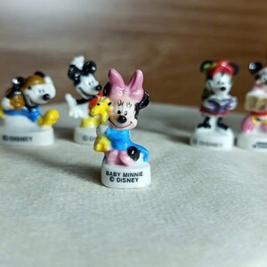 Puede incluir: Una colecci&oacute;n de peque&ntilde;as figuras de Disney, incluyendo a Baby Minnie Mouse, Mickey Mouse y otros personajes. Las figuras est&aacute;n sobre bases blancas y presentan colores vibrantes. Baby Minnie, en primer plano, lleva un lazo rosa y un vestido azul.
