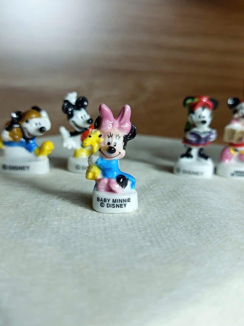 Puede incluir: Una colecci&oacute;n de peque&ntilde;as figuras de Disney, incluyendo a Baby Minnie Mouse, Mickey Mouse y otros personajes. Las figuras est&aacute;n sobre bases blancas y presentan colores vibrantes. Baby Minnie, en primer plano, lleva un lazo rosa y un vestido azul.