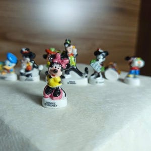 以下が含まれることがあります： ミニーマウス、ドナルドダック、グーフィーなど、小さなディズニーキャラクターのフィギュアのコレクション。ミニーマウスは、黄色のドレスとピンクのリボンを着て、前景にいます。フィギュアは白い表面にあります。