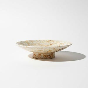 May include: A speckled ceramic pedestal bowl. The bowl has a wide, shallow dish with a speckled cream and brown pattern. It sits on a short, round pedestal base with a similar speckled design. The bowl is likely used for serving or display.