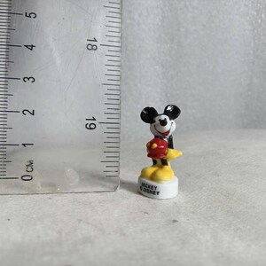 May include: A small Mickey Mouse figurine, approximately 1 inch tall, stands on a white base. Mickey is depicted in his classic attire: black ears, a red jacket, yellow shoes, and white gloves. The base reads "Mickey Disney."