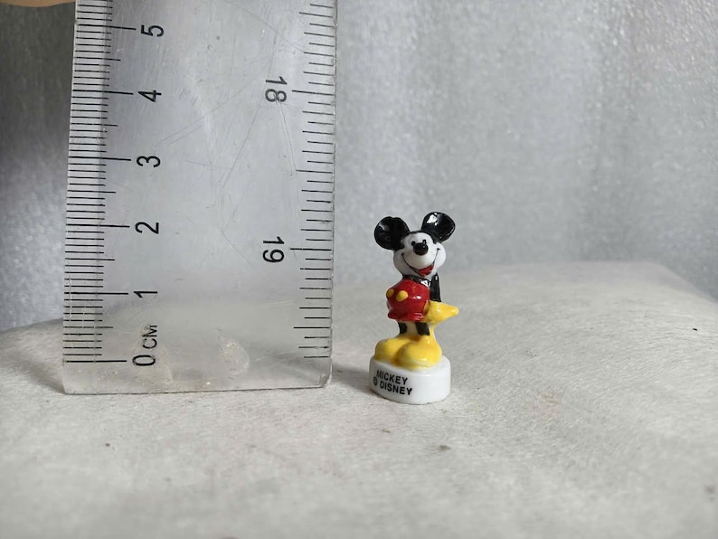 May include: A small Mickey Mouse figurine, approximately 1 inch tall, stands on a white base. Mickey is depicted in his classic attire: black ears, a red jacket, yellow shoes, and white gloves. The base reads "Mickey Disney."