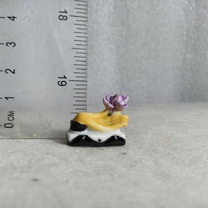 May include: A miniature ceramic sculpture featuring a yellow hand holding a purple flower. The sculpture is mounted on a black and white base. The sculpture is approximately 1 inch tall.