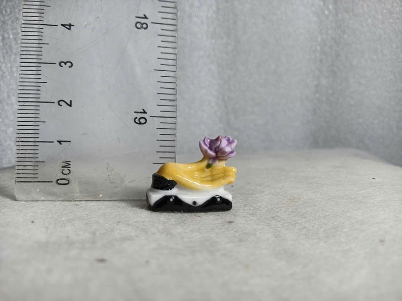 May include: A miniature ceramic sculpture featuring a yellow hand holding a purple flower. The sculpture is mounted on a black and white base. The sculpture is approximately 1 inch tall.