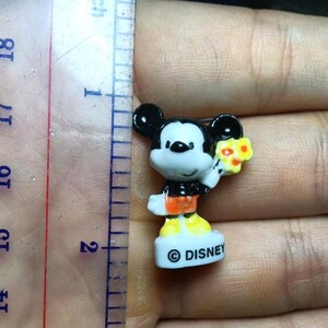 Op de afbeelding: Een kleine Mickey Mouse-figuur, ongeveer 2 cm hoog, met een boeket gele bloemen. Het beeldje draagt een zwarte broek, een oranje shirt en gele schoenen. De basis is wit met het "&copy; DISNEY" logo.