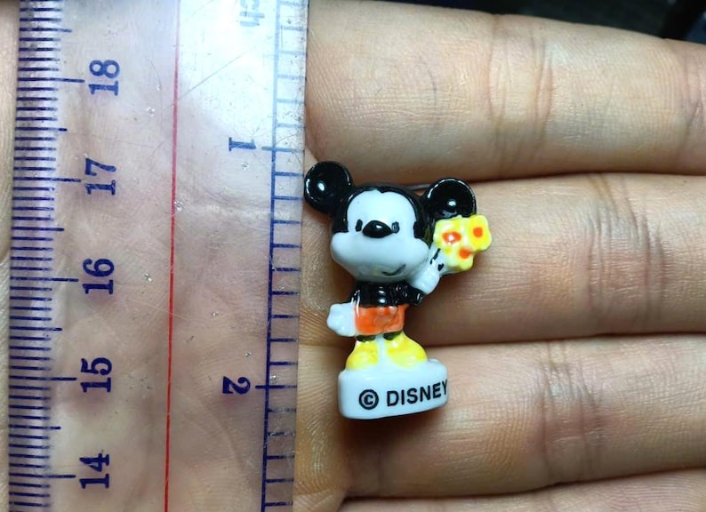 Op de afbeelding: Een kleine Mickey Mouse-figuur, ongeveer 2 cm hoog, met een boeket gele bloemen. Het beeldje draagt een zwarte broek, een oranje shirt en gele schoenen. De basis is wit met het "&copy; DISNEY" logo.
