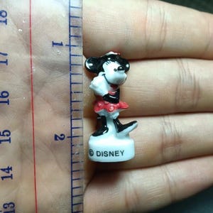 May include: A small Minnie Mouse figurine, approximately 1.5 inches tall, with a black and white head, red and white dress, and black shoes. The figurine is standing on a white base with the word "DISNEY" printed on it.