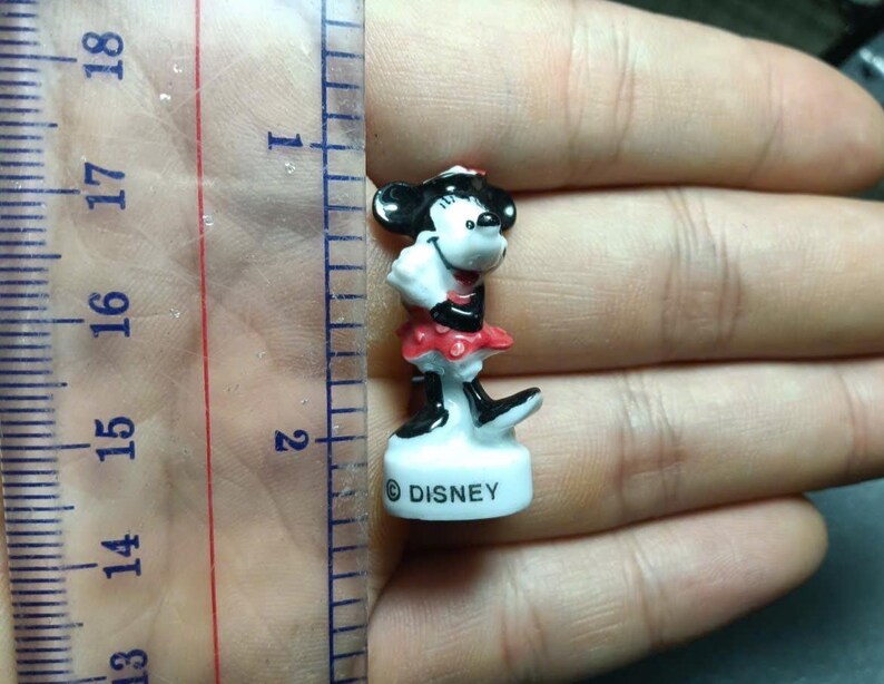 May include: A small Minnie Mouse figurine, approximately 1.5 inches tall, with a black and white head, red and white dress, and black shoes. The figurine is standing on a white base with the word "DISNEY" printed on it.