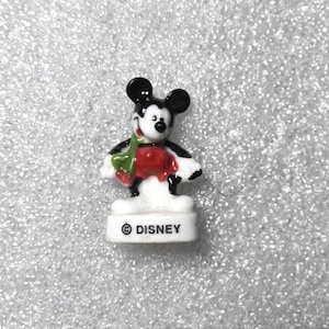 May include: A small Mickey Mouse figurine with black ears, a black body, and a red shirt. The figurine is holding a green object and stands on a white base with the Disney logo.