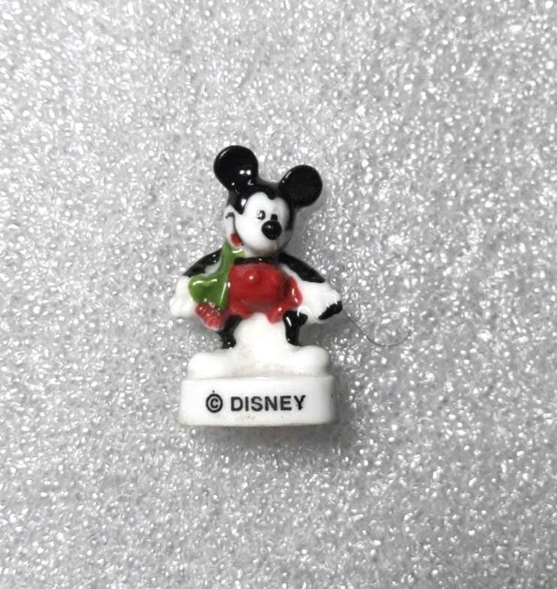 May include: A small Mickey Mouse figurine with black ears, a black body, and a red shirt. The figurine is holding a green object and stands on a white base with the Disney logo.