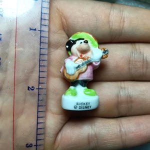 Puede incluir: Peque&ntilde;a figura de Mickey Mouse, vestido con un atuendo colorido y tocando la guitarra. La figura mide aproximadamente 5 cm de alto y tiene una base blanca con el texto "Mickey Disney". La figura se sostiene en una mano.
