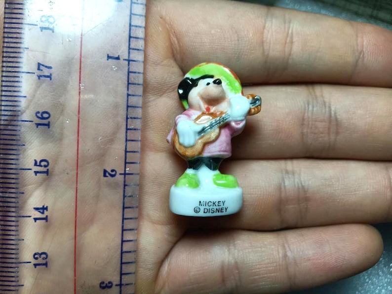 Puede incluir: Peque&ntilde;a figura de Mickey Mouse, vestido con un atuendo colorido y tocando la guitarra. La figura mide aproximadamente 5 cm de alto y tiene una base blanca con el texto "Mickey Disney". La figura se sostiene en una mano.
