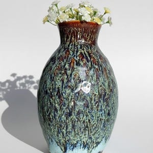 May include: A ceramic vase with a textured, mottled glaze in shades of teal, brown, and blue. The vase has a wide body and a narrow neck, and is filled with small white flowers. The vase is approximately 8 inches tall.