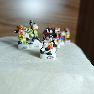以下が含まれることがあります： 小さなディズニーキャラクターのフィギュアのコレクション。ミッキーマウスは、黒いシルクハットとタキシードを着て前面にいます。他のキャラクターには、ドナルドダック、グーフィー、ミニーマウスが含まれます。各フィギュアは、ディズニーのロゴが付いた白いベースにあります。