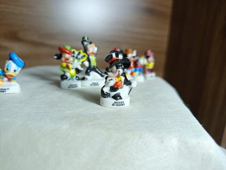以下が含まれることがあります： 小さなディズニーキャラクターのフィギュアのコレクション。ミッキーマウスは、黒いシルクハットとタキシードを着て前面にいます。他のキャラクターには、ドナルドダック、グーフィー、ミニーマウスが含まれます。各フィギュアは、ディズニーのロゴが付いた白いベースにあります。