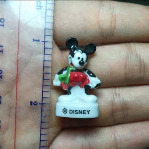 May include: A small Mickey Mouse figurine, approximately 1 inch tall, in a classic pose. The figure is white, black, red, and green, with the Disney logo on the base. The figurine is held in a hand.