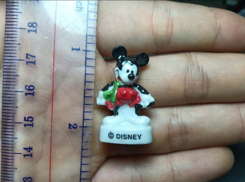 May include: A small Mickey Mouse figurine, approximately 1 inch tall, in a classic pose. The figure is white, black, red, and green, with the Disney logo on the base. The figurine is held in a hand.