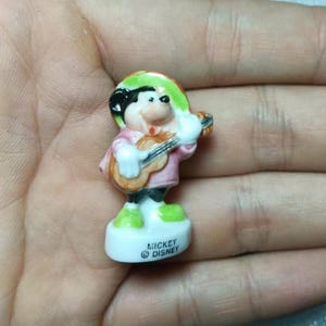 Puede incluir: Peque&ntilde;a figura de cer&aacute;mica de Mickey Mouse tocando la guitarra. Mickey viste camisa rosa, sombrero verde y pantal&oacute;n negro. La figura est&aacute; sobre una base blanca con el texto "Mickey Disney".