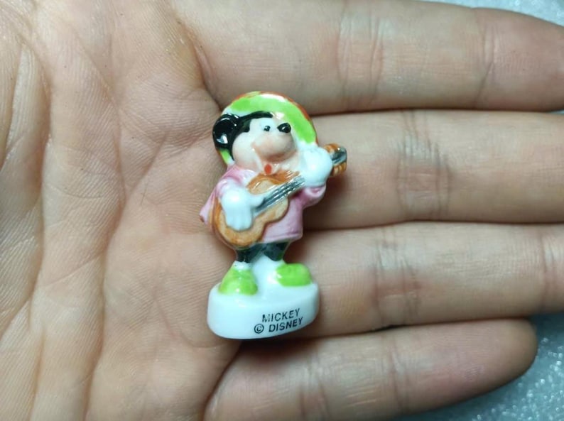 Puede incluir: Peque&ntilde;a figura de cer&aacute;mica de Mickey Mouse tocando la guitarra. Mickey viste camisa rosa, sombrero verde y pantal&oacute;n negro. La figura est&aacute; sobre una base blanca con el texto "Mickey Disney".