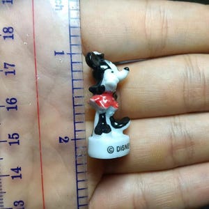 May include: A small Minnie Mouse figurine, approximately 1.5 inches tall, in a classic pose. The figure is black, white, and red, with the Disney logo on the base. The figurine is held in a hand next to a ruler.