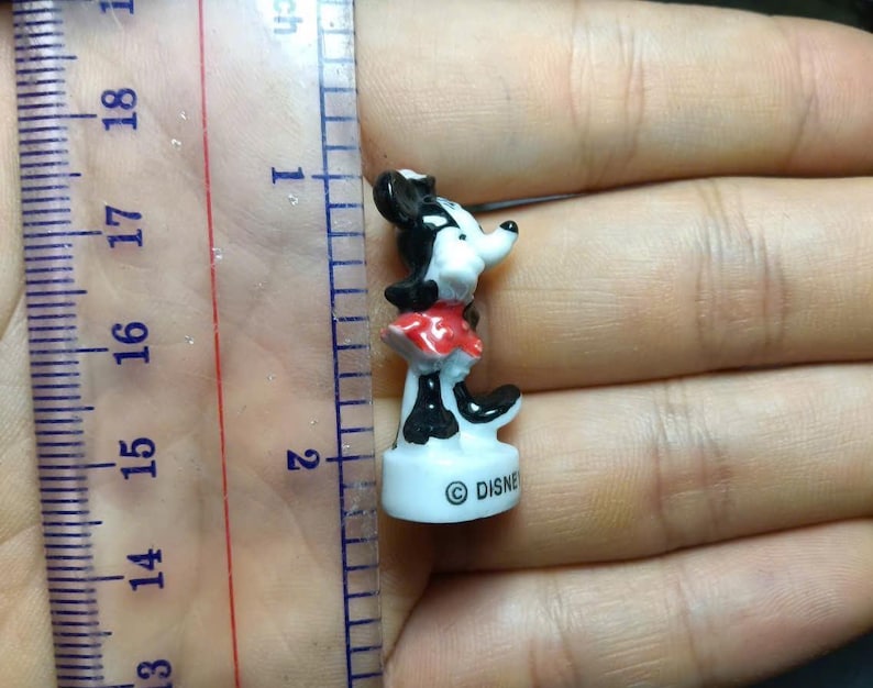 May include: A small Minnie Mouse figurine, approximately 1.5 inches tall, in a classic pose. The figure is black, white, and red, with the Disney logo on the base. The figurine is held in a hand next to a ruler.
