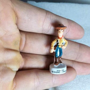 Pu&ograve; includere: Una piccola statuina di Woody del franchise Disney Pixar Toy Story. La statuina indossa un cappello da cowboy marrone, una camicia gialla con pois neri, pantaloni blu e stivali marroni. La base &egrave; bianca con la scritta "WOODY DISNEY/PIXAR".