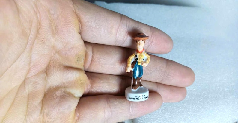 Pu&ograve; includere: Una piccola statuina di Woody del franchise Disney Pixar Toy Story. La statuina indossa un cappello da cowboy marrone, una camicia gialla con pois neri, pantaloni blu e stivali marroni. La base &egrave; bianca con la scritta "WOODY DISNEY/PIXAR".