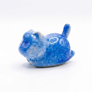 Könnte beinhalten: Kleine Katzenfigur aus Keramik in Blautönen. Die Katze liegt mit erhobenem Schwanz. Die Figur hat ein strukturiertes, kristallines Aussehen mit Variationen in der blauen Farbe. Ein dekorativer Artikel.