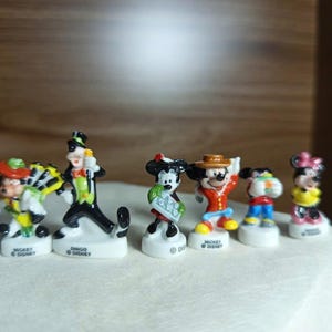 以下が含まれることがあります： 小さくてカラフルなディズニーキャラクターのフィギュアのコレクション。ミッキーマウス、ミニーマウス、グーフィーなど、それぞれユニークな衣装とポーズをしています。白い台座に置かれ、ディズニーのロゴが見えます。