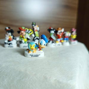 以下が含まれることがあります： ミッキーマウス、ミニーマウス、ドナルドダックなど、小さなディズニーキャラクターのフィギュアのコレクション。フィギュアは明るい色で、白い台座に立っています。「BABY DONALD DISNEY」の文字が台座に表示されています。