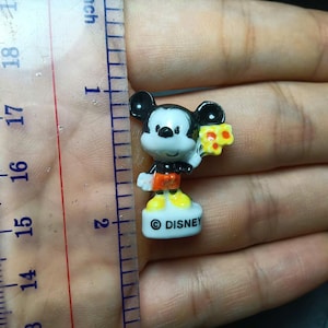 May include: A miniature Mickey Mouse figure, about 2.5 cm tall, holding a piece of cheese. The figure features black ears, a white face, red shorts, and yellow shoes. The white base is inscribed with the word "DISNEY".
