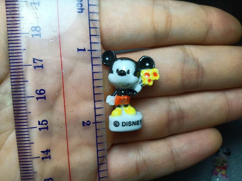 May include: A miniature Mickey Mouse figure, about 2.5 cm tall, holding a piece of cheese. The figure features black ears, a white face, red shorts, and yellow shoes. The white base is inscribed with the word "DISNEY".