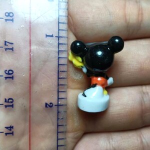Op de afbeelding: Een kleine Mickey Mouse-figuur, ongeveer 4 cm hoog, met een zwarte kop en oren, een oranje broek en witte handschoenen en schoenen. De figuur houdt een geel voorwerp vast. De figuur staat op een witte basis.