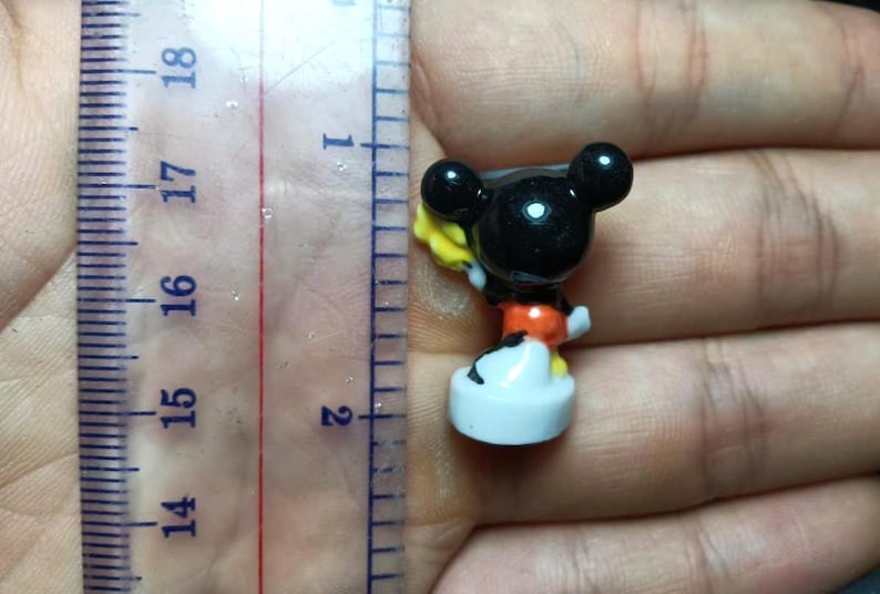 Op de afbeelding: Een kleine Mickey Mouse-figuur, ongeveer 4 cm hoog, met een zwarte kop en oren, een oranje broek en witte handschoenen en schoenen. De figuur houdt een geel voorwerp vast. De figuur staat op een witte basis.