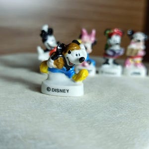 Puede incluir: Colecci&oacute;n de figuras de personajes de Disney. La figura del primer plano es Mickey Mouse, con sombrero marr&oacute;n, chaqueta azul y zapatos amarillos. La base de la figura es blanca con el logo de Disney. Otros personajes son visibles en el fondo.