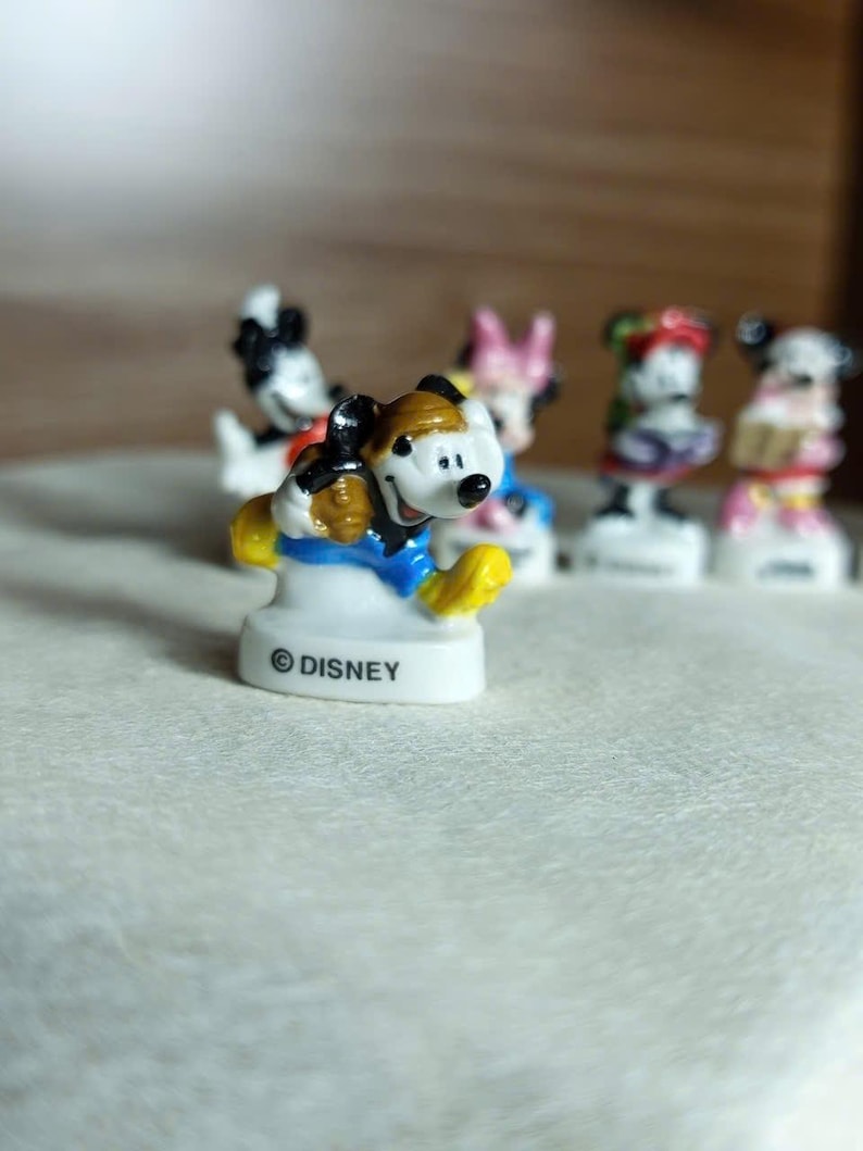 Puede incluir: Colecci&oacute;n de figuras de personajes de Disney. La figura del primer plano es Mickey Mouse, con sombrero marr&oacute;n, chaqueta azul y zapatos amarillos. La base de la figura es blanca con el logo de Disney. Otros personajes son visibles en el fondo.