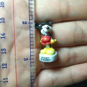 May include: A miniature Mickey Mouse figure, about 3 cm tall, is positioned near a ruler. The figure features black ears, a white face, red shorts, yellow shoes, and a black jacket. The base is inscribed with "Mickey &copy; Disney."