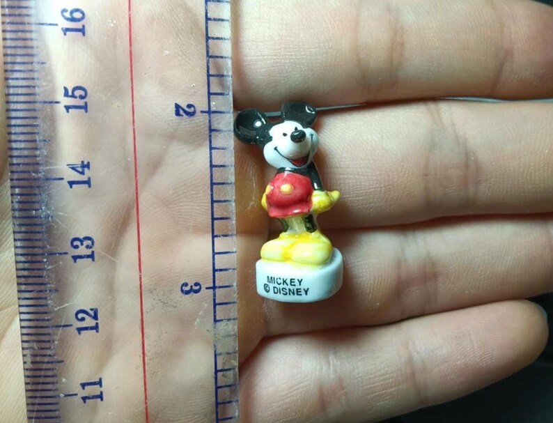 May include: A miniature Mickey Mouse figure, about 3 cm tall, is positioned near a ruler. The figure features black ears, a white face, red shorts, yellow shoes, and a black jacket. The base is inscribed with "Mickey &copy; Disney."
