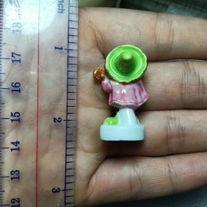 Puede incluir: Peque&ntilde;a figura pintada a mano de una persona con un vestido rosa y un sombrero verde. La figura mide aproximadamente 3,8 cm de altura y se apoya sobre una base blanca. Sostiene un objeto peque&ntilde;o.