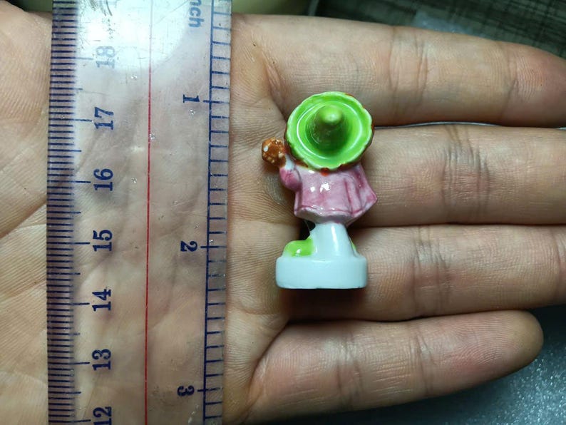 Puede incluir: Peque&ntilde;a figura pintada a mano de una persona con un vestido rosa y un sombrero verde. La figura mide aproximadamente 3,8 cm de altura y se apoya sobre una base blanca. Sostiene un objeto peque&ntilde;o.