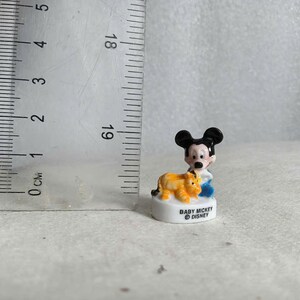 May include: A small ceramic figurine of Baby Mickey Mouse with a yellow and orange lion. Mickey is wearing a blue outfit. The base is white with the text "BABY MICKEY &copy; DISNEY". The figurine is approximately 1 inch tall.