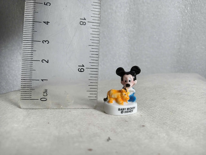 May include: A small ceramic figurine of Baby Mickey Mouse with a yellow and orange lion. Mickey is wearing a blue outfit. The base is white with the text "BABY MICKEY &copy; DISNEY". The figurine is approximately 1 inch tall.