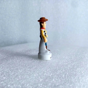 Pu&ograve; includere: Una piccola statuina del personaggio Woody del film Toy Story. La statuina indossa un cappello da cowboy marrone, una camicia gialla e pantaloni blu. La base &egrave; bianca.