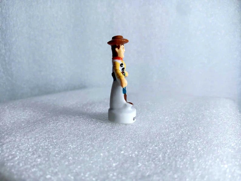 Pu&ograve; includere: Una piccola statuina del personaggio Woody del film Toy Story. La statuina indossa un cappello da cowboy marrone, una camicia gialla e pantaloni blu. La base &egrave; bianca.